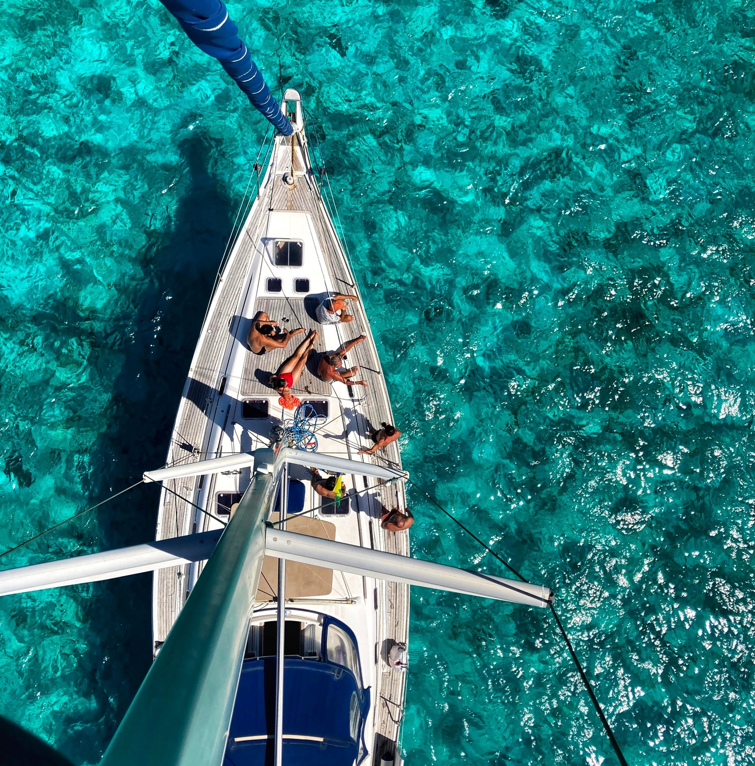 A photo taken from above, the top of the sailboat’s mass. It shows a group of friends laying on the deck, with crystal clear blue water around them.
