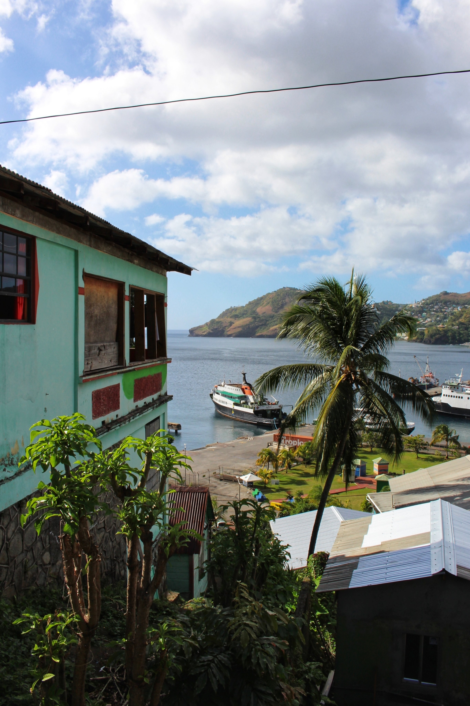 A typical Caribbean picture showing a colorful house on the left, palm tree on the right with the sea, a ship and the rest of the harbor clearly visible in the background.